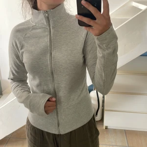 Ljusgrå ziptröja med hög krage - Säljer en ljusgrå ziptröja med lång ärm och hög krage. Tröjan har dragkedja hela vägen och är i ett mjukt, stretchigt material. Sitter inte åt i handlarna. Mjukt fluffigt material inuti. Från Nelly. Använd en gång. Stl M men passar S.