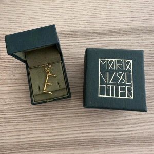Guldhalsband med bokstav E från Maria Nilsdotter - Snyggt halsband i guld från Maria Nilsdotter med ett hänge i form av bokstaven E. Kedjan är tunn och elegant, och bokstaven har en stilren design. Perfekt för dig som vill bära din initial eller ge bort som personlig present.