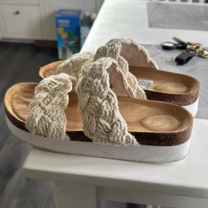 Beige flätade sandaler med platåsula - Säljer ett par beige sandaler från Chic med två breda, flätade band i grovstickat material. Skorna har en bekväm platåsula i korkimitation och vit undersida. Perfekta för varma dagar och ger en avslappnad men trendig look. Använda 3 gånger kanske så är i jätte fint skick! 