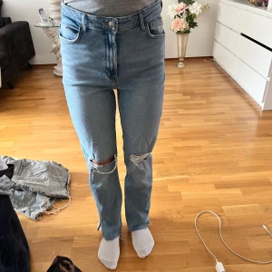 Ljusblå högmidjade jeans med hål på knäna - Säljer ett par ljusblå jeans med hög midja och raka ben. Jeansen har slitna detaljer och hål på båda knäna för en trendig look. Klassisk femficksmodell med knapp och dragkedja framtill.