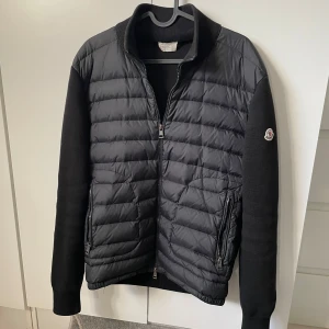 Moncler cardigan - Snygg svart jacka från Moncler med quiltad framsida och stickade ärmar. Jackan har dragkedja framtill, två fickor med dragkedja och ribbad krage. Klassisk Moncler-logga på ärmen. Perfekt för kyliga dagar. Den är i jätte bra skick o säljer endast för den inte kommer till användning storleken är M men den hade passat s lika bra😊