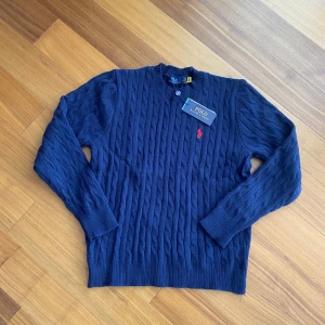 Polo Ralph Lauren Flätstickad Marinblå Tröja – Helt Ny med Taggar (Storlek L) - Säljer en helt ny flätstickad tröja från Polo Ralph Lauren i marinblått – aldrig använd, alla taggar sitter kvar. Klassisk design med röd broderad logotyp på bröstet. Tröjan är i storlek L och passar perfekt både till vardags och mer uppklätt.  ✔️ Storlek: L ✔️ Skick: Ny, med taggar ✔️ Färg: Marinblå ✔️ Material: 100% bomull ✔️ Äkta vara 📦 Skickas spårbart eller kan mötas upp i Stockholm 💬 Pris kan diskuteras vid snabb affär 