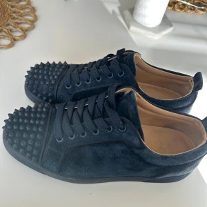 Mörkblå sneakers från Christian Louboutin - Säljer ett par mörkblå sneakers i mocka från Christian Louboutin med röda sulor och mörkblå nitar på tån. Skorna har snörning och rund tå, samt läderfoder. Skorna är lätt slitna men i väldigt bra skick. Storlek 43. 42 fransk storlek