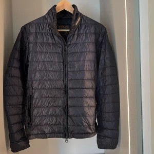  Woolrich jacka - Snygg blå quiltad lättviktsjacka från Woolrich med dragkedja framtill och två fickor. Jackan har en klassisk passform och är perfekt för kyliga dagar. Tunn och smidig att bära, med diskret logga i nacken. Finns en liten defekt på bild 4!!!