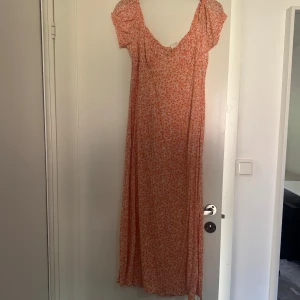 Blommig långklänning med puffärmar - Långklänning i ljus orange med små vita blommor och korta puffärmar. Klänningen har v-ringning och smockdetaljer vid bysten.