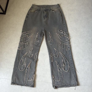 Grå jeans med råa broderade detaljer - Unika grå jeans med bred passform och råa, broderade mönster längs benen. Snygga slitna kanter och coola detaljer både fram och bak. Perfekta för dig som vill sticka ut med din stil.