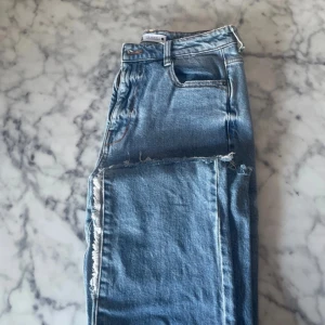 Blå raka jeans med råa kanter - Säljer ett par blå jeans med slitna detaljer längs benen. Hög midja och dragkedja. Använda ett fåtal gånger.
