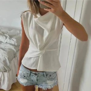 Vit omlottopp med knyt - Säljer en snygg vit omlottopp med knyt i midjan. Toppen har ärmlös design och en draperad effekt framtill som ger en cool och modern look. Perfekt att styla med jeansshorts eller kjol.