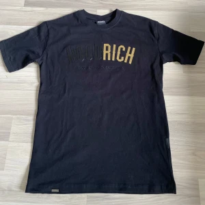 Svart t-shirt från Hoodrich - Svart t-shirt från Hoodrich med tryckt logga i svart och guld på bröstet. Klassisk rund halsringning och korta ärmar. Tillverkad i mjuk bomull för en bekväm känsla.
