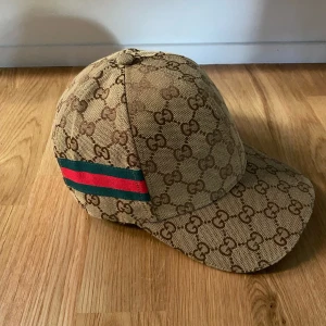 Beige keps från Gucci med monogram - Säljer en beige keps från Gucci med klassiskt GG-monogram över hela kepsen och grönt/rött band på sidan. Justerbar rem baktill i brunt skinn. Perfekt accessoar för en trendig look.