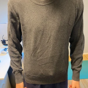 Grå stickad tröja från J.Lindeberg - Säljer en stilren grå stickad tröja från J.Lindeberg. Tröjan har rund halsringning och ett diskret strukturerat mönster. Perfekt för lager-på-lager och passar till många olika outfits.