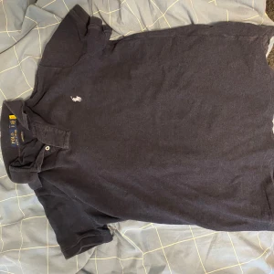 Mörkblå pikétröja från Polo Ralph Lauren - Säljer en mörkblå pikétröja från Polo Ralph Lauren med klassisk krage och två knappar framtill. Tröjan har en broderad logga på bröstet och är i modellen custom slim fit. Perfekt för en stilren och avslappnad look. Säljer eftersom den är för liten