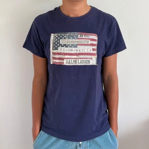 Ralph lauren denim&supply flag T-shirt  - Snygg mörkblå t-shirt från Ralph Lauren Denim & Supply med coolt amerikanskt flaggtryck och vintagekänsla på bröstet. Klassisk passform och rund halsringning, tillverkad i mjuk bomull. Perfekt för en avslappnad och stilren look.