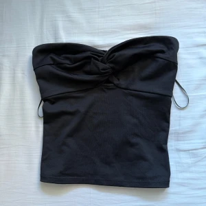 Svart bandeau tubtopp med twist - Säljer en svart bandeau tubtopp med snygg twistdetalj framtill och smockad baksida för extra stretch. Toppen är axelbandslös och har en clean, stilren look som passar perfekt till höga jeans eller kjol. Materialet känns mjukt och följsamt. Ge en väldigt fin form och sitter bra
