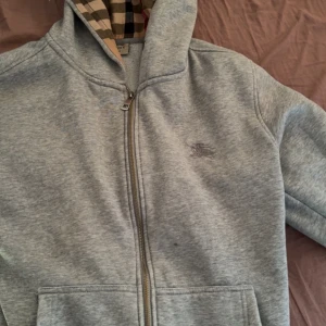 Grå hoodie från Burberry med dragkedja - Grå hoodie från Burberry i bomull med klassiskt rutigt foder i huvan. Tröjan har hel dragkedja, fickor framtill och broderad logga på bröstet. Perfekt för dig som gillar stilrena och lyxiga streetwear-vibbar.