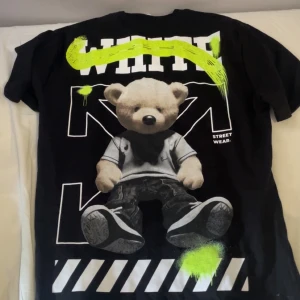 Svart Off-White t-shirt med nalletryck - Svart t-shirt från Off-White med stort tryck av en nalle på ryggen och mindre nalletryck fram. Grafiska detaljer i vitt och neon-gult, samt texten 'STREET WEAR.' Trycket har street-art känsla och coola färgklickar. Perfekt för dig som gillar unika statement-plagg.