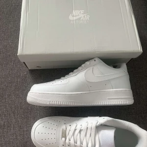 Nike Air Force 1 - Klassiska Nike Air Force 1 sneakers i helvitt skinn med perforerad tå, vit sula och ikonisk swoosh på sidan. Lågt skaft, vita skosnören och diskret silverdetalj framtill. Perfekta för en clean och stilren look. För liten för mig och fick dem som present.