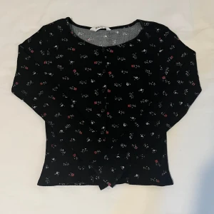 Svart blommig kofta från H&M - Supersöt svart kofta från H&M med små blommor i vitt och rött. Koftan har knappar framtill, rund halsringning och trekvartsärm. Materialet är ribbat och stretchigt, vilket gör den både bekväm och snygg till jeans eller kjol.