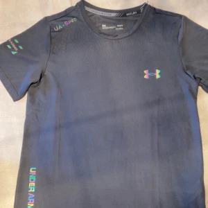 Svart Under Armour t-shirt för träning - Sportig svart t-shirt från Under Armour med tryck på bröstet och ärmen. Gjord i lätt och snabbtorkande polyester, perfekt för löpning eller gym. Snygga detaljer med logga och text i reflekterande färg. Klassisk passform och rund hals. Pris kan diskuteras.