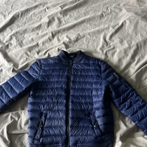 Blå pufferjacka från Polo Ralph Lauren - Snygg marinblå pufferjacka från Polo Ralph Lauren med quiltad design, dragkedja framtill och ribbade muddar. Jackan har en patch med logga på ärmen och praktiska fickor med dragkedja. Perfekt för kyliga dagar och riktigt clean stil. 