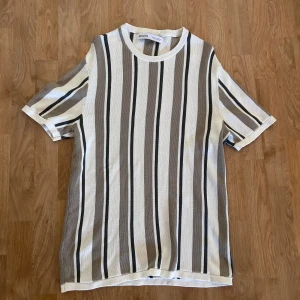 Randig t-shirt från Bershka - Snygg randig t-shirt från Bershka i beige, grått, svart och vitt. Modellen har korta ärmar, rund halsringning och är tillverkad i ett mjukt bomullstyg. 