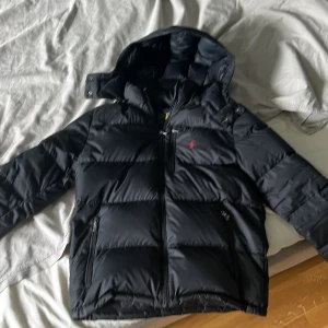 Svart pufferjacka från Polo Ralph Lauren - Snygg svart pufferjacka från Polo Ralph Lauren med röd broderad logga på bröstet. Jackan har huva, dubbla dragkedjor och flera praktiska fickor. Perfekt för kalla dagar och riktigt skön att bära. Klassisk och stilren design som funkar till allt. Storlek XL i barn som kan bäras som S i vuxen storlek. Kan bäras från 165cm-180. Lägg gärna prisförslag. Inga defekter, repor eller hål,