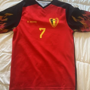 Belgien De Bruyne #7 fotbollströja - Snygg Belgien fotbollströja med De Bruyne och nummer 7 i gult tryck. Röd bas med svarta partier och coola eldliknande detaljer på ärmarna. V-ringad hals och korta ärmar. Perfekt för dig som vill visa support för landslaget.