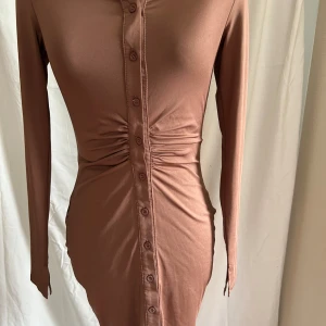 Brun figurnära klänning med knappar - Snygg brun kortklänning från ZY i stretchigt material med långa ärmar och knappar hela vägen framtill. Klänningen har rynkade detaljer vid midjan som ger en cool siluett. 