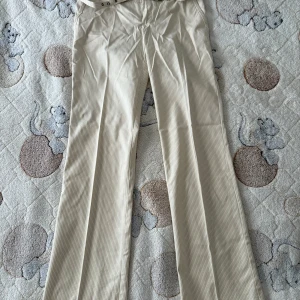 Beige randiga kostymbyxor från Mexx - Snygga beige kostymbyxor från Mexx med diskreta ränder. Byxorna har klassisk rak passform och pressveck framtill för en clean look. Perfekta för dig som gillar stilrena och tidlösa plagg. Materialet är mjukt och bekvämt, passar till många outfits.