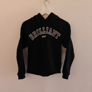 Lindex hoodie - Svart hoodie från Lindex med texten 'BRILLIANT no1' i vitt framtill. Tröjan har huva, långa ärmar och ribbade muddar. Tillverkad i mjuk bomull, perfekt för en chill och avslappnad stil.