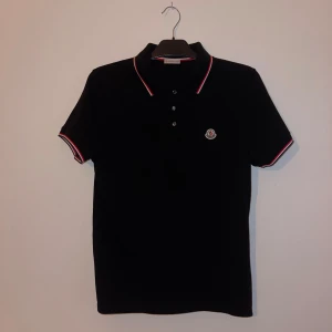 Svart Moncler pikétröja med röd/vit kant - Svart pikétröja från Moncler med korta ärmar och klassisk krage. Röd och vit randig kant på ärmslut och krage. 