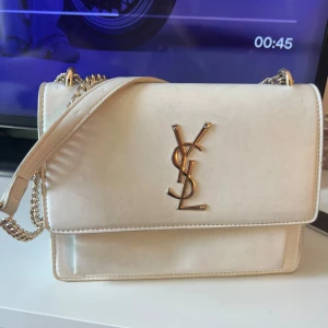 Vit axelväska från Yves Saint Laurent - !inte riktig! Snygg vit axelväska från YSL i skinn med guldiga kedjedetaljer och ikoniskt YSL-emblem framtill. Väskan har ett stilrent, fyrkantigt format och flera fack inuti för smart organisering. Perfekt för dig som vill ha en lyxig och klassisk accessoar.