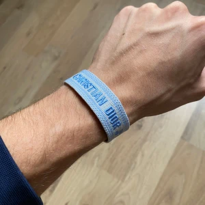 Christian Dior blått tygarmband - Snyggt armband från Christian Dior i ljusblått tyg med texten 'CHRISTIAN DIOR J'ADIOR' broderad i blått. Armbandet har en enkel och stilren design med justerbar knytning och fransar i ändarna. Perfekt accessoar för att lyfta din outfit.