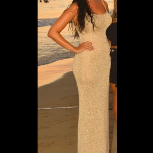 Glittrig champagnefärgad långklänning - Supersnygg långklänning i champagnefärg med glittrigt tyg och smala axelband. Klänningen har en tight passform och en hög slits på sidan som ger en extra touch. Perfekt för dig som vill sticka ut med en elegant och trendig look.