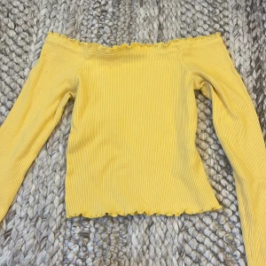 Gul offshoulder topp från Gina Tricot - Gul offshoulder topp från Gina Tricot med långa ärmar. Toppen är ribbad och har volangkant upptill och nertill. Storlek M men materialet är stretchigt och passar mindre storlekar. 