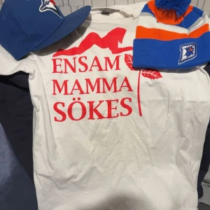 Vit t-shirt med röd text - Vit t-shirt med trycket 'ENSAM MAMMA SÖKES' i rött och en röd siluett ovanför texten. Klassisk passform och rund halsringning. Perfekt statement-plagg för dig som vill sticka ut.