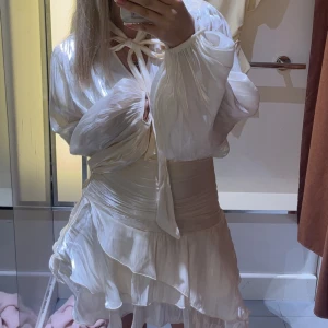 Mini frill dress - Superfin kort klänning i champagnefärgat, créme vit glansigt material med långa puffärmar, omlottliv och knytband i halsen. 