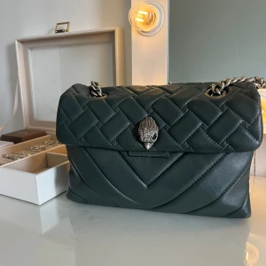 Quiltad axelväska från Kurt Geiger - Snygg mörkgrön quiltad axelväska från Kurt Geiger med silverfärgad kedjerem och ikonisk fågeldekor på locket. Väskan är tillverkad i mjukt skinn och har magnetlås. Perfekt storlek för det viktigaste och ger en lyxig känsla till din outfit.