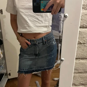 Blå jeanskjol med fransig kant - Säljer en snygg blå jeanskjol med rå, fransig nederkant och klassiska fickor både fram och bak. Kjolen har en kort passform och stängs med knapp och dragkedja framtill. Perfekt för dig som gillar en avslappnad och trendig stil.