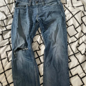 Blå Levis 506 jeans med slitning - Klassiska blå Levis 506 jeans med rak passform och snyggt sliten look. Jeansen har en tydlig reva på vänster ben för en extra edge. Perfekta för dig som gillar en avslappnad och tidlös stil.