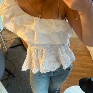 Vit omlott topp med volang och broderi - Superfin vit topp med omlott-design och stora volanger över ena axeln. Toppen är ärmlös och har broderade detaljer med små hål, vilket ger en romantisk känsla. Perfekt till jeans eller kjol för en fräsch sommarlook.