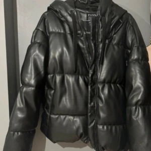 Svart pufferjacka i skinnimitation - Säljer en svart pufferjacka från Panda med glansig skinnimitation, huva med snörning och dragkedja framtill. Jackan är quiltad och har en oversized vibe, perfekt för kalla dagar. Snygg och trendig look som lyfter vilken outfit som helst.