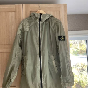 Ljusgrön jacka från Stone Island - Säljer en ljusgrön jacka från Stone Island med huva och dragkedja framtill. Jackan har två snedställda fickor och den klassiska Stone Island-loggan på vänster ärm. Tillverkad i lätt nylonmaterial, perfekt för blåsiga dagar. Passar som M-L.