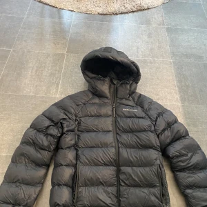 Svart dunjacka Peak Performance XS - Säljer en svart dunjacka från Peak Performance i storlek XS. Jackan har huva, dragkedja framtill och två sidofickor med dragkedja. Perfekt för kalla dagar och riktigt skön att bära. Klassisk pufferstil med diskret logga på bröstet.
