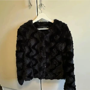 Svart pälsjacka från Vero Moda - Säljer en svart pälsjacka från Vero Moda med coolt vågigt mönster i pälsen. Jackan är fluffig och har rund halsringning samt knäppning framtill. Perfekt för dig som vill sticka ut med en trendig och mysig look.