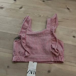 Randig rosa croppad topp från Zara - Superfin croppad topp från Zara i rosa och vitt med smala ränder. Toppen har breda axelband med volangdetaljer och resår i midjan för en snygg passform. Perfekt till sommaren och lätt att matcha med jeans eller kjol.