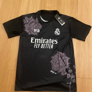 Real Madrid x Y-3 svart fotbollströja M - Snygg svart Real Madrid fotbollströja från Adidas Y-3 och Yohji Yamamoto. Tröjan har korta ärmar, vita tryck med klubbmärke och sponsor samt unika blommiga detaljer. Tillverkad i lätt funktionsmaterial (AEROREADY) som andas. Perfekt för dig som vill sticka ut på planen.