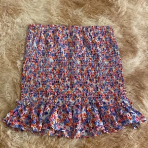 Blommig volangkjol från FB Sister - Supersöt kort kjol från FB Sister med blommigt mönster i blått, rosa, orange och vitt. Kjolen har smockad midja och en volang längst ner som ger en lekfull vibe. Perfekt för dig som gillar färg och vill sticka ut lite extra.