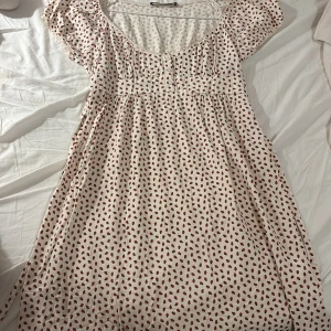 Vit kortklänning med jordgubbsmönster - Supersöt vit kortklänning från Brandy Melville med röda jordgubbar över hela tyget. Klänningen har korta ärmar, smock i midjan och knappar framtill. Perfekt för sommarens alla häng och picknickar. Lätt och flowy modell som ger en chill vibe. Köpt i London nu i år, aldrig använt utan endast provat hemma men tycker tyvärr den har lite för djup u-ringning för min smak! Men perfekt skick annars!