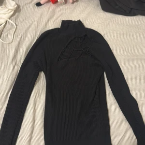 Svart ribbad polotröja med snörning - Svart ribbad polotröja med lång ärm och cool snörningsdetalj över bröstet. Tröjan är tight i modellen och har hög krage. Perfekt för dig som gillar en edgy och trendig look.